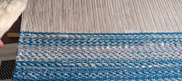in progress weaving; the bottom has woven weft in varying blue colors, the top has unwoven warp that is white. a fluff of white is shown on the left.