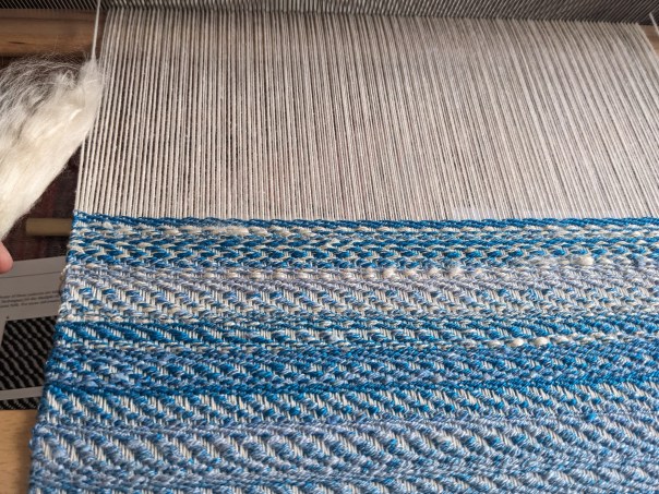 in progress weaving; the bottom has woven weft in varying blue colors, the top has unwoven warp that is white. a fluff of white is shown on the left.