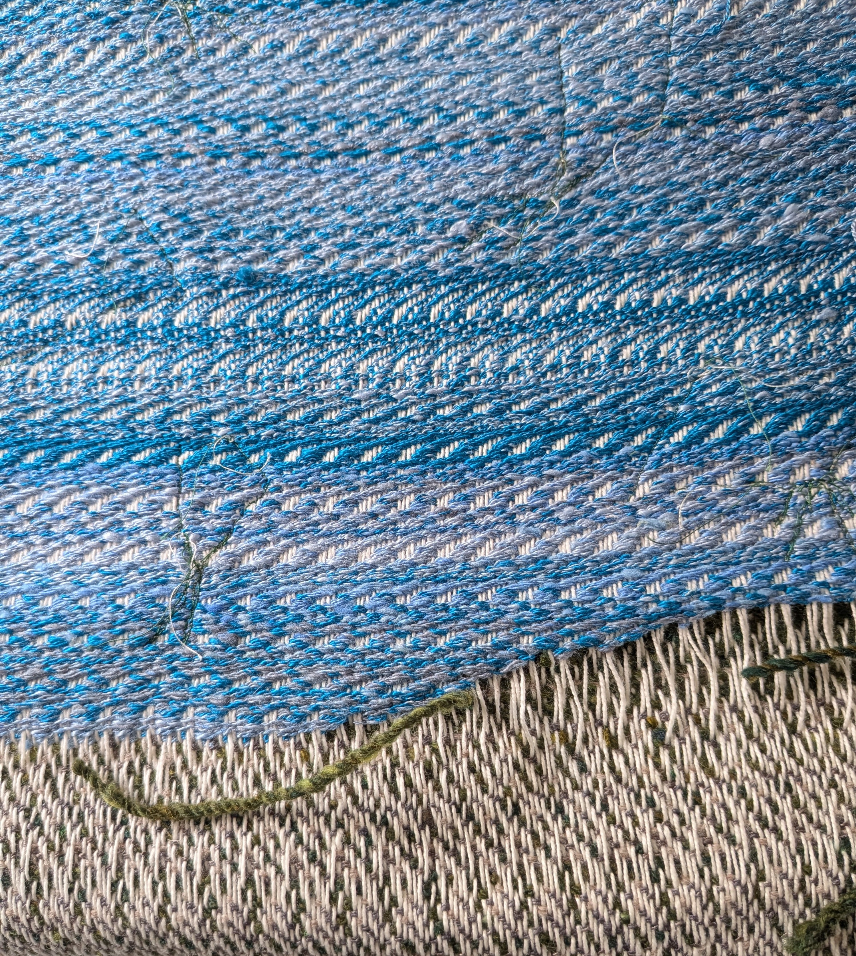 handwoven fabric, bottom is white with spots of green and the top is stripes of blue and white. some yarn is sticking out