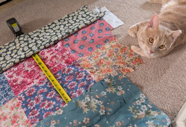 on the left, a bunch of floral fabrics with a measuring tape. a cat on the right looks towards the camera